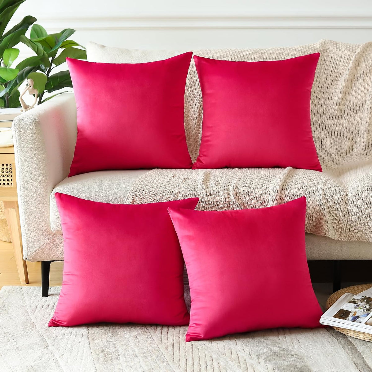 Four bright fuchsia square velvet pillows staged on a cream sofa with a knit throw and magazine nearby.