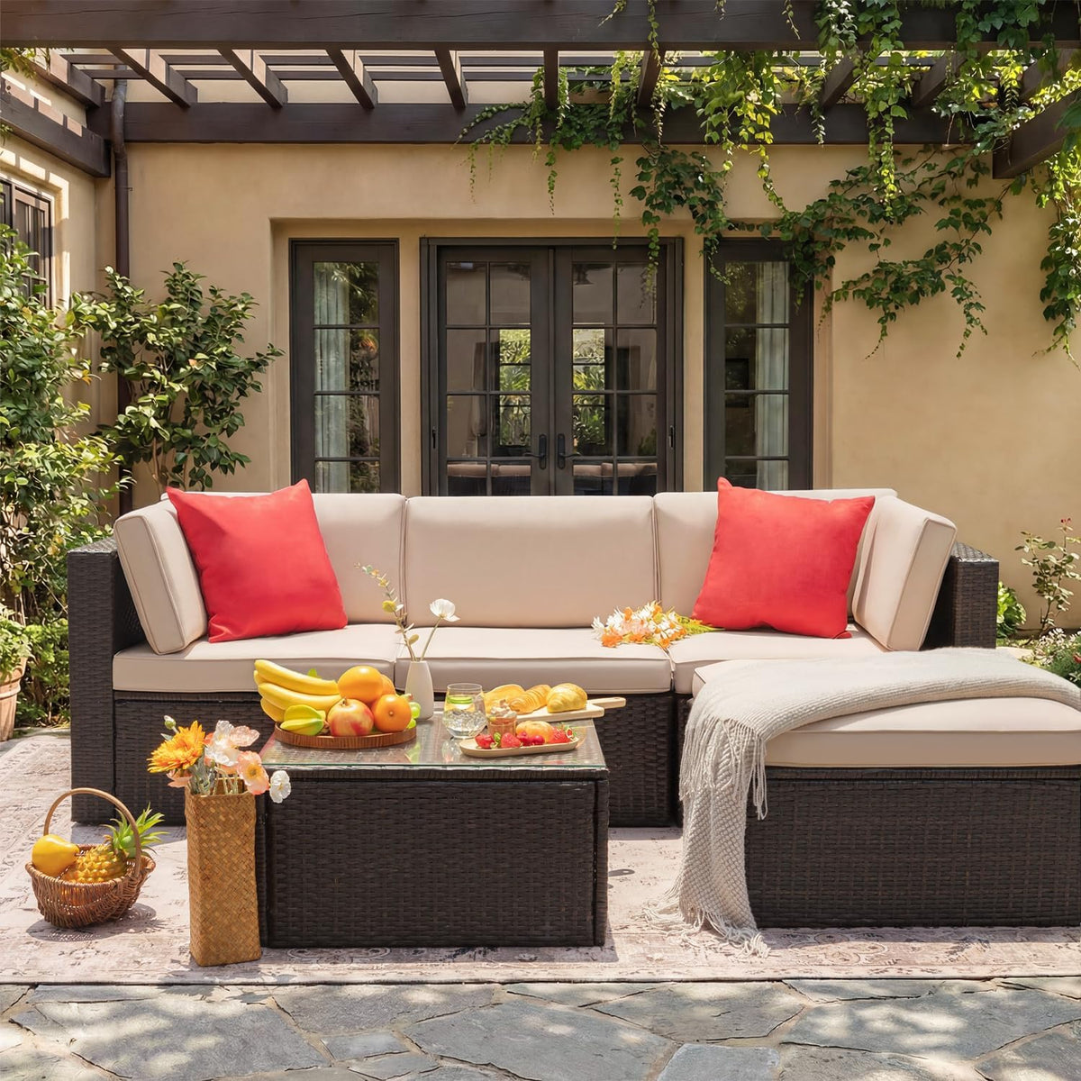Front view of the beige wicker sectional under a pergola with red pillows, throw blanket, and a coffee table