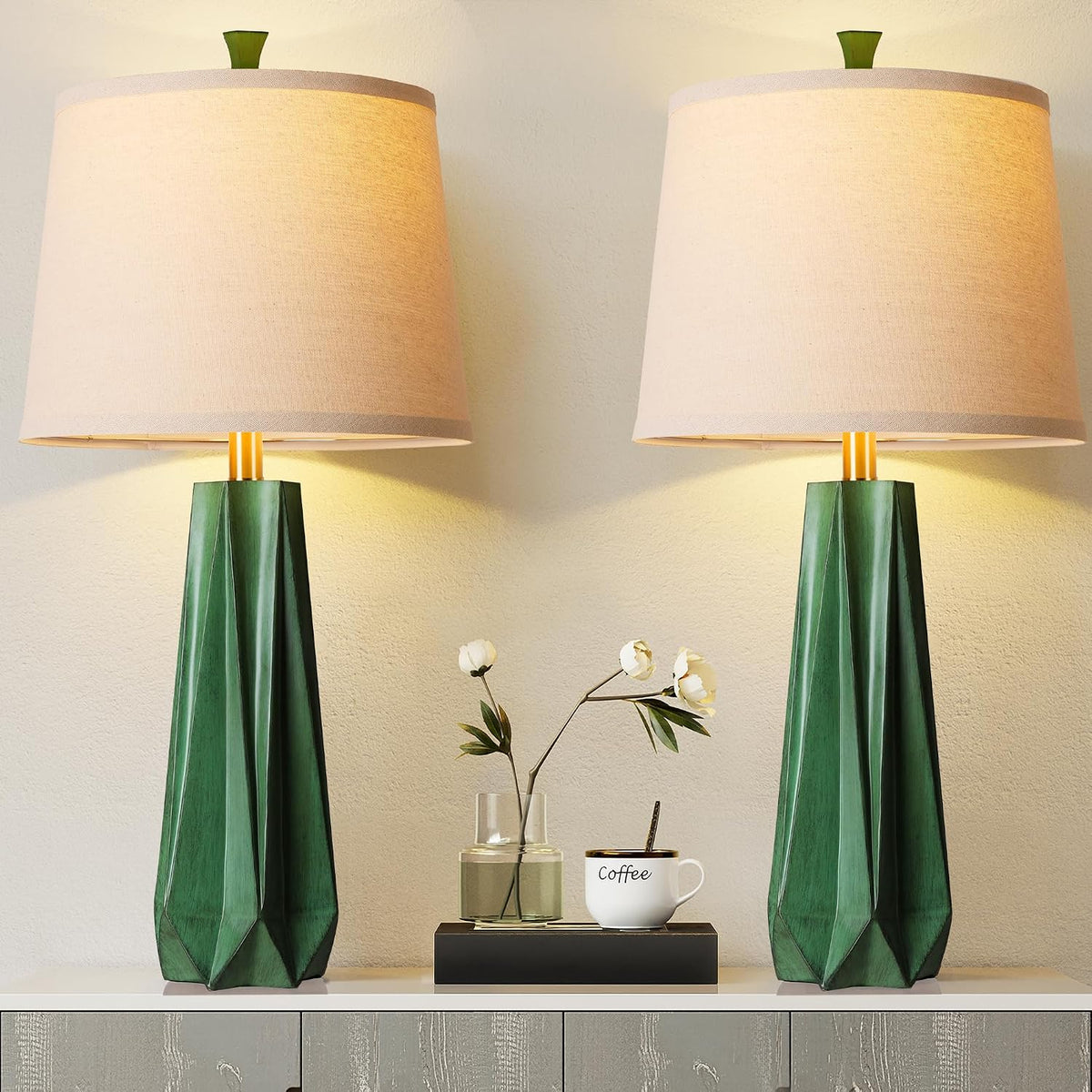 Front view of two green-finished geometric table lamps with beige linen shades flanking a small vase and coffee cup.