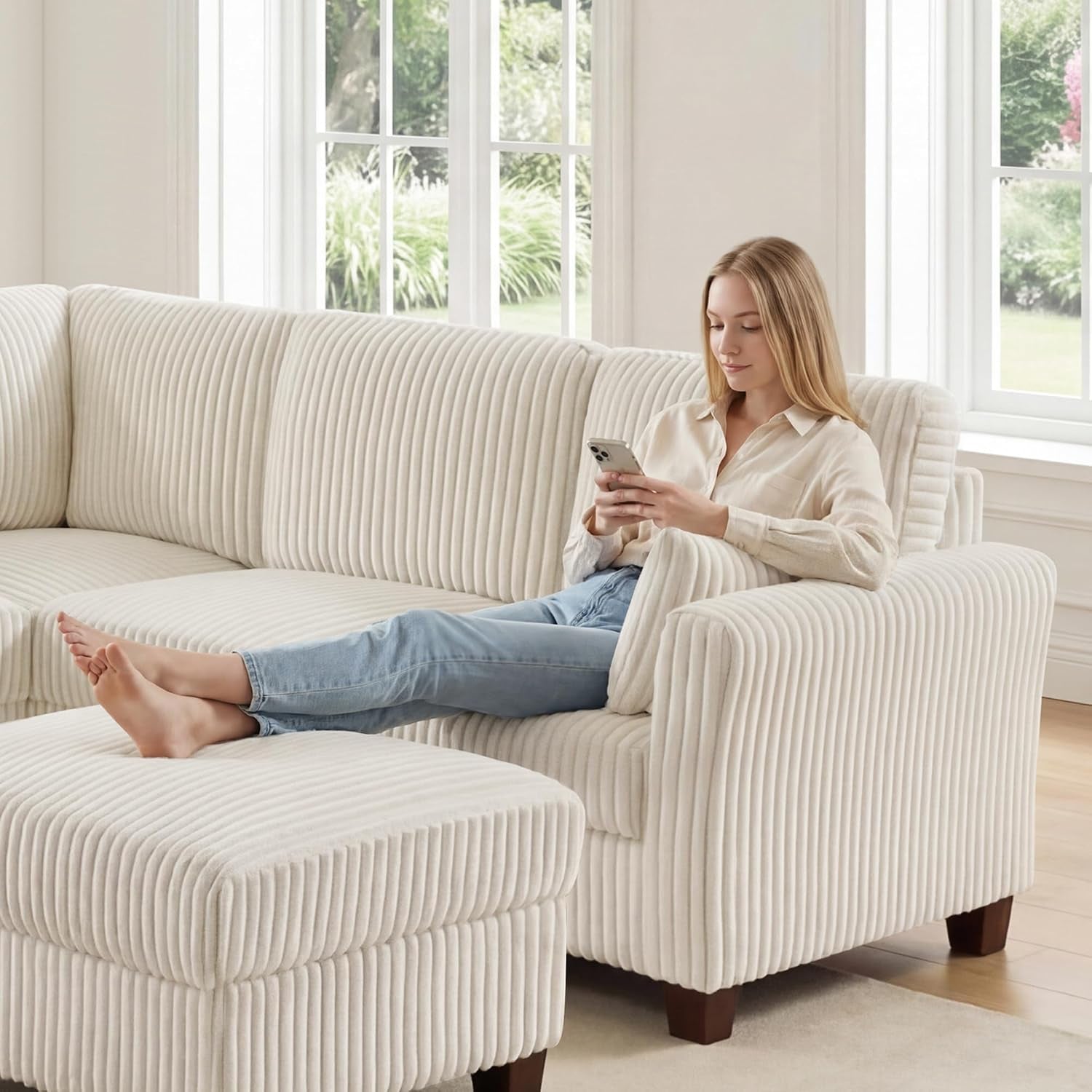 Woman reclining on a cream corduroy modular corner sectional with matching ottoman by sunlit windows.