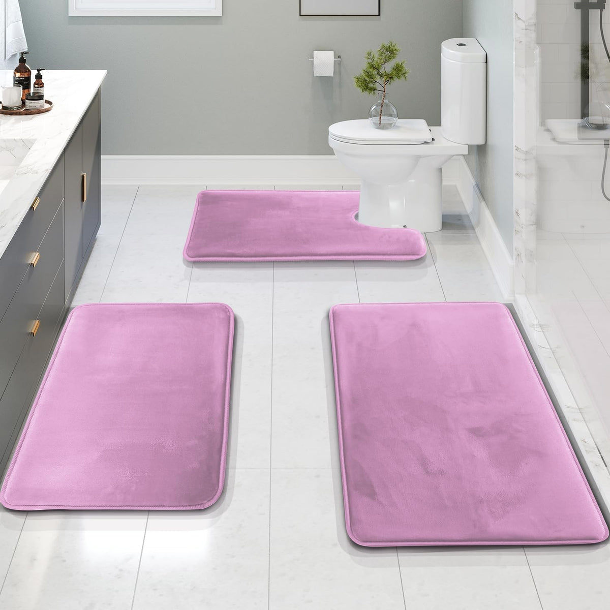 Low-angle bathroom view with three lavender-pink memory foam rugs leading toward a U-shaped contour mat by the toilet.