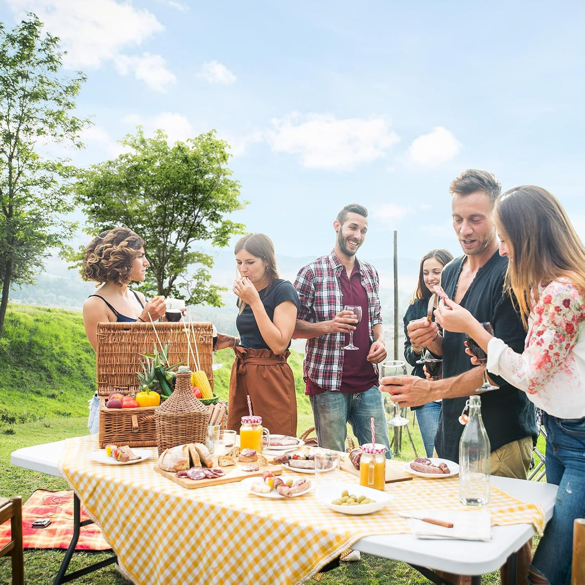 Outdoor picnic with a white folding table topped by a yellow check tablecloth and food while people socialize.