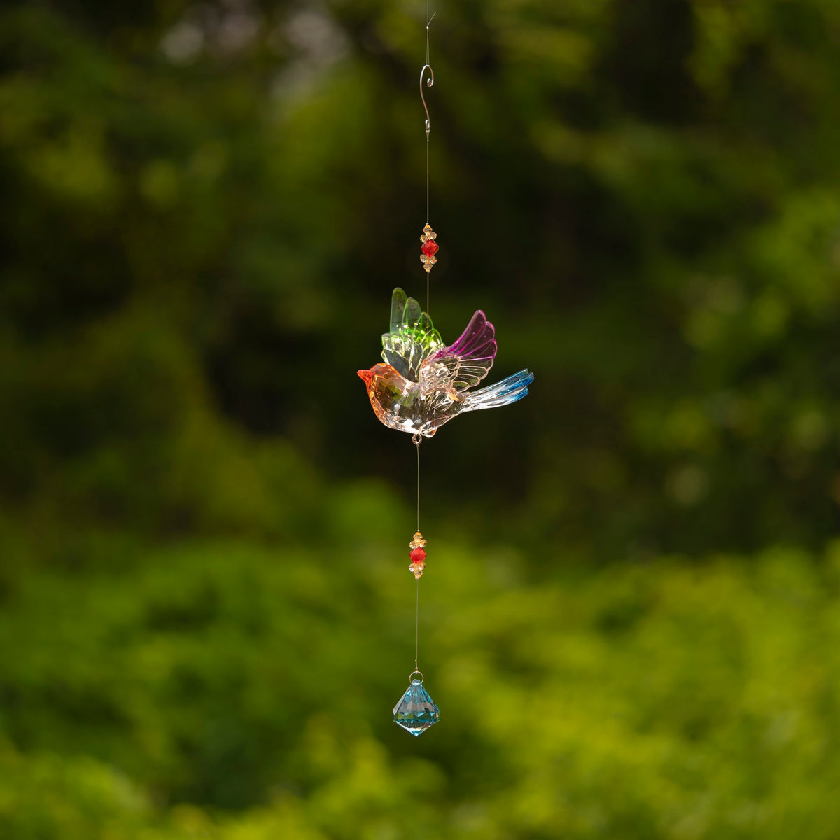A small multicolored bird ornament hangs from a long string against a leafy green background.
