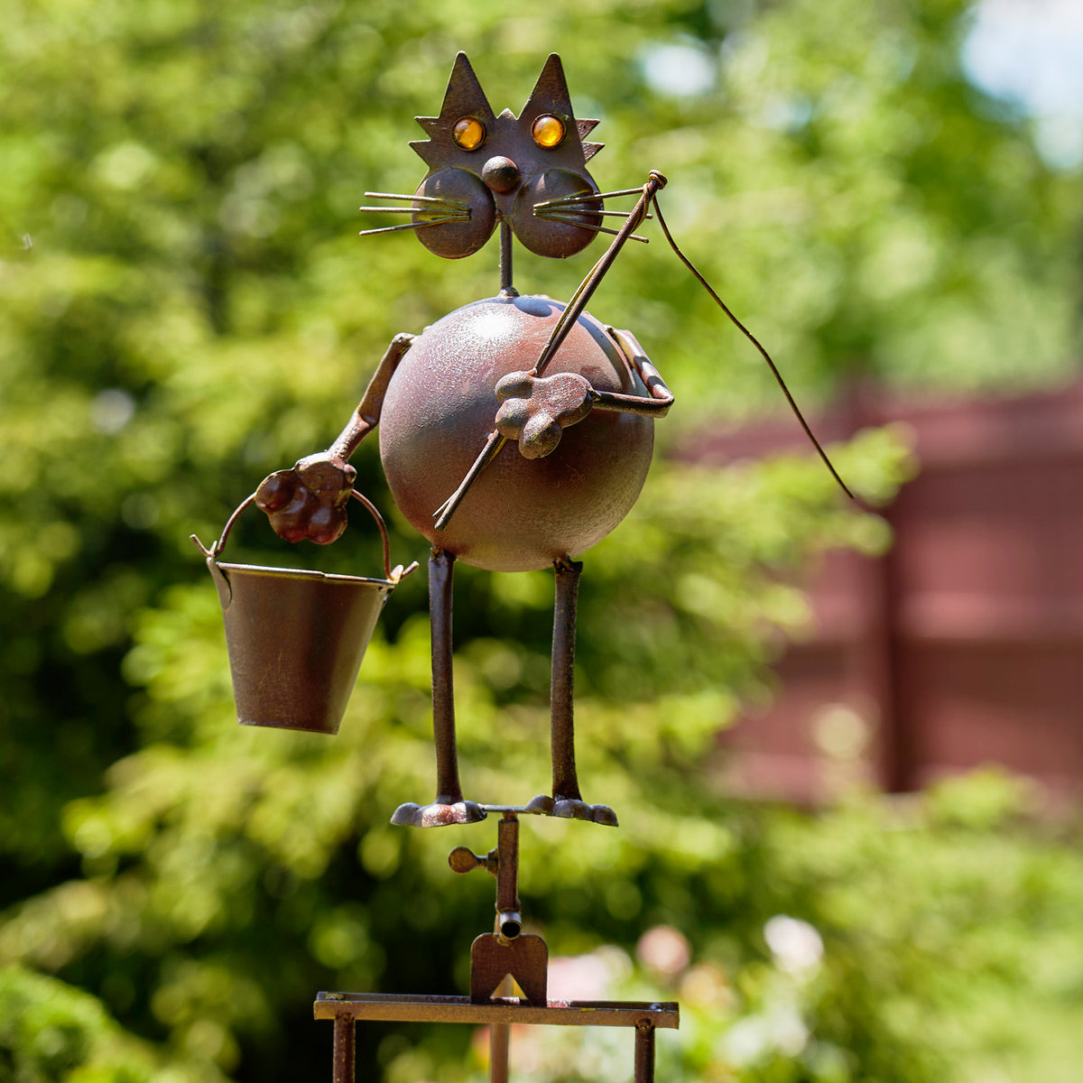 Rust-colored metal cat garden stake holding a fishing rod and bucket, with a round glass ball body.