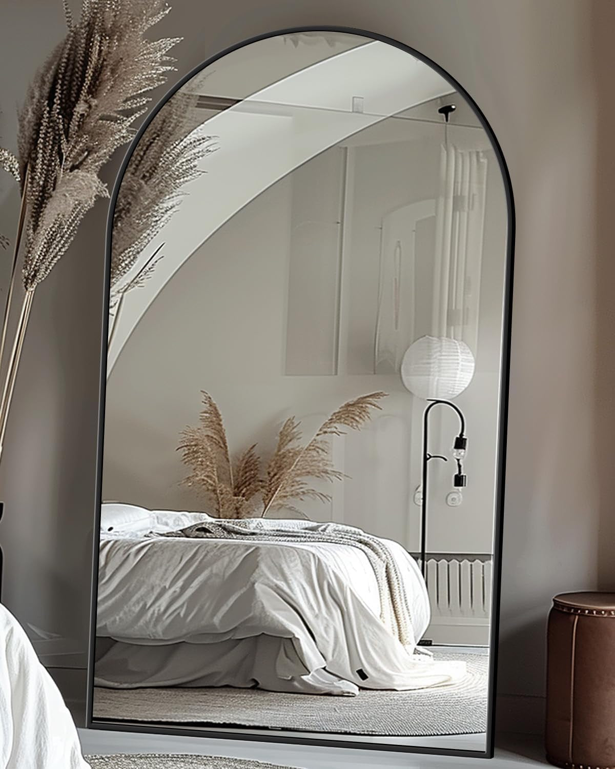Arched full-length mirror with slim black frame standing on floor, reflecting a bed and pampas grass