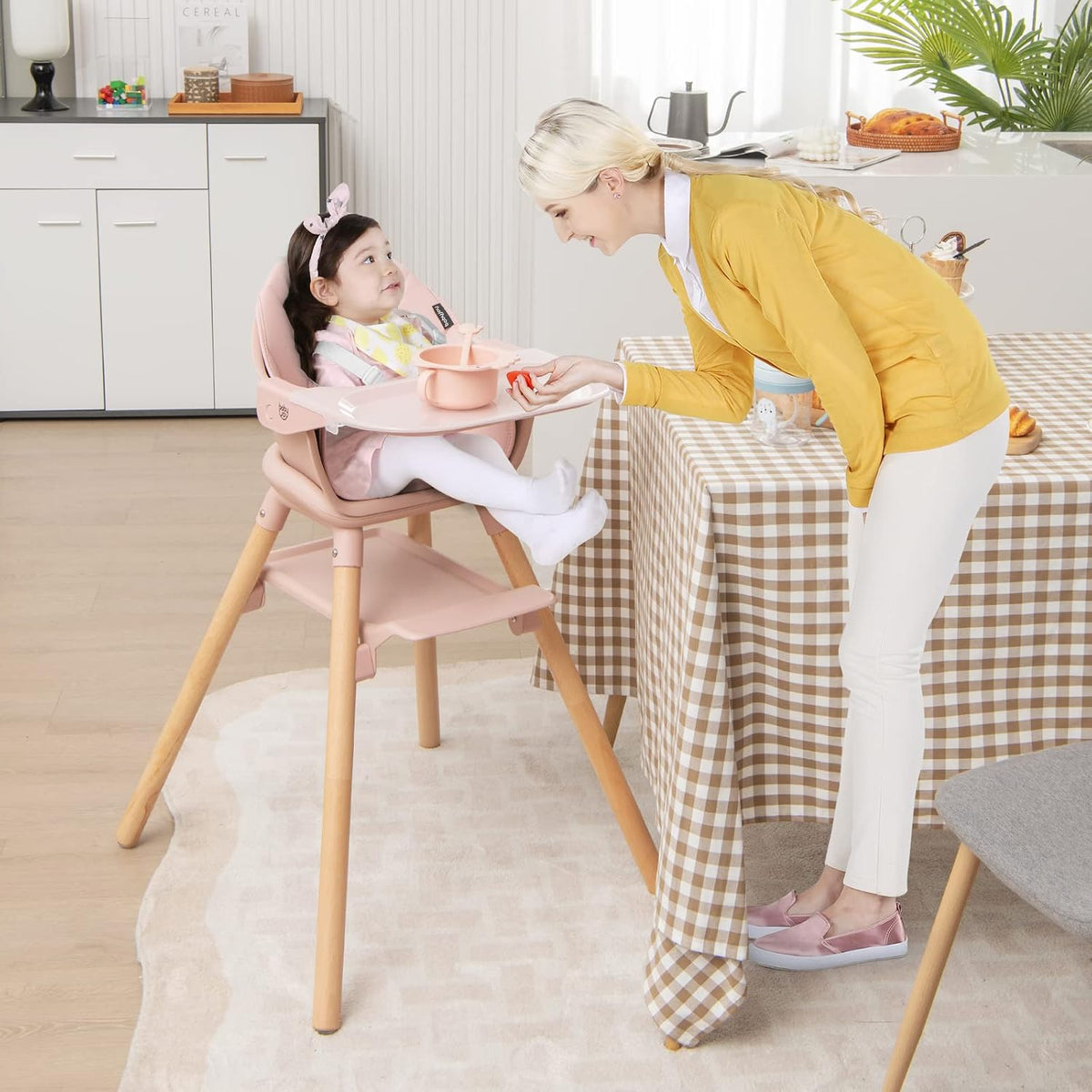 A toddler seated in the pink high chair at a table while an adult leans over to feed them.