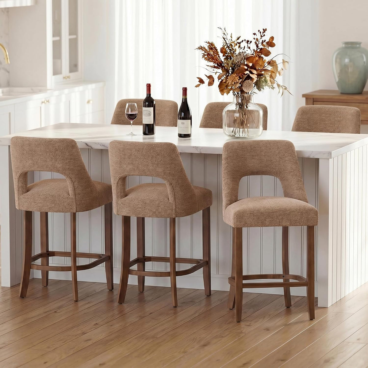 Three sand-toned textured counter stools with curved back openings and warm wood legs lined at a white island, a wine bottle