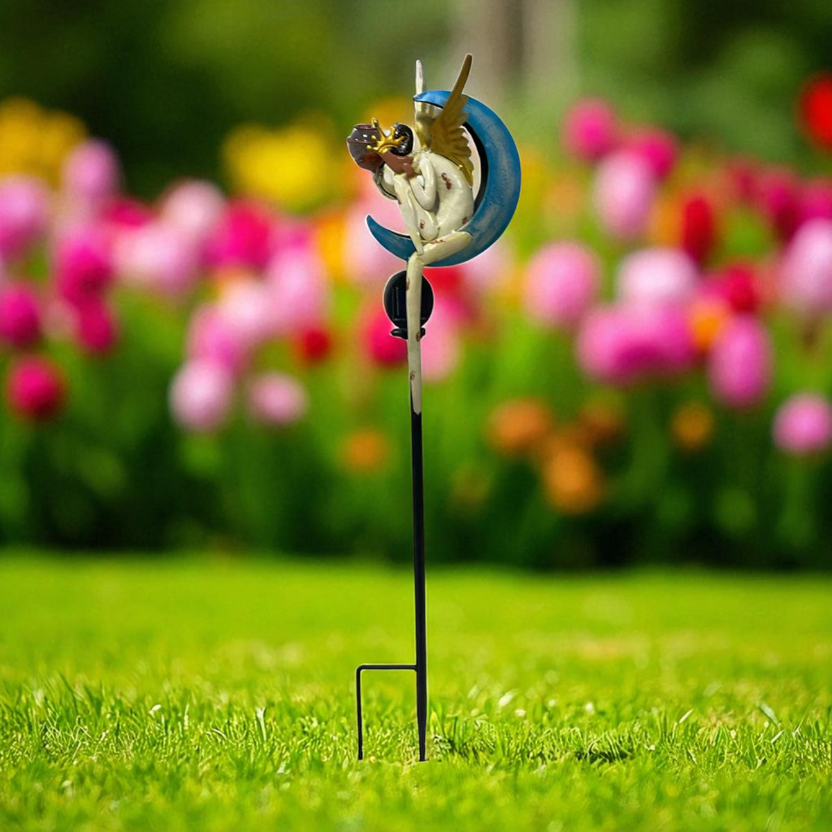 Side view of an angel seated against a blue crescent, mounted on a slender stake in a flower garden.