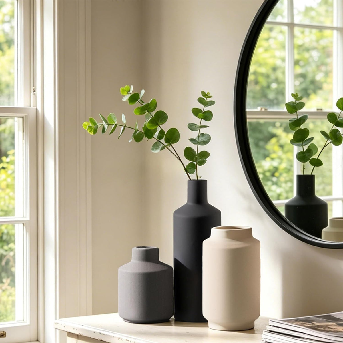 Tall black matte vase with eucalyptus stems on a dresser near a round mirror and window reflection