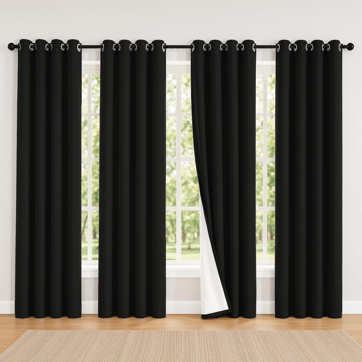 Wide window dressed with four black grommet curtains, one panel pulled back to show white backing and wood floor rug below.