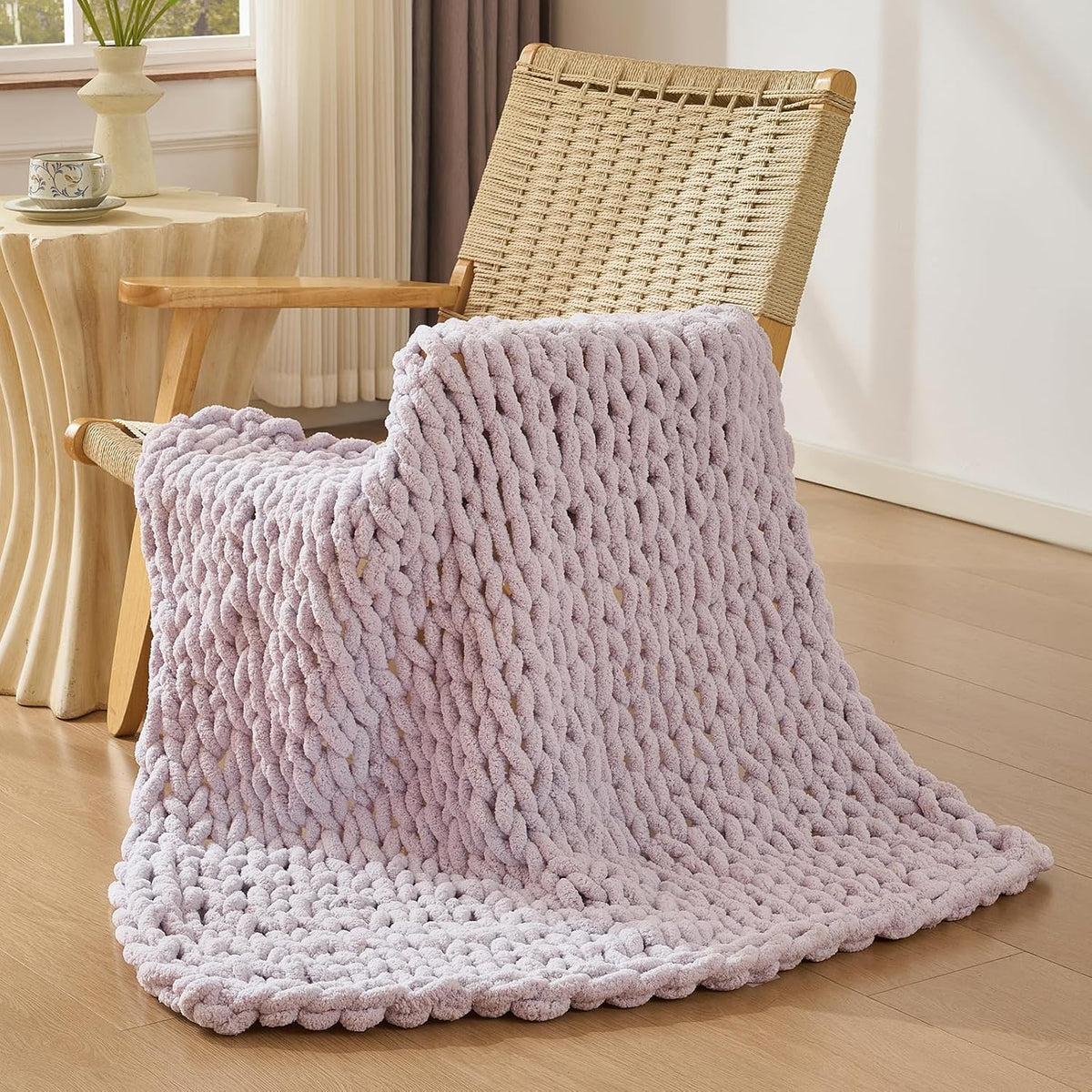 Soft lavender chunky knit throw draped over the seat and arm of a woven wooden chair in a bright room.