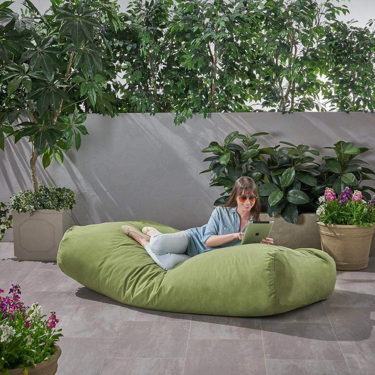 Woman reclining on green floor lounger on a tiled patio surrounded by potted plants