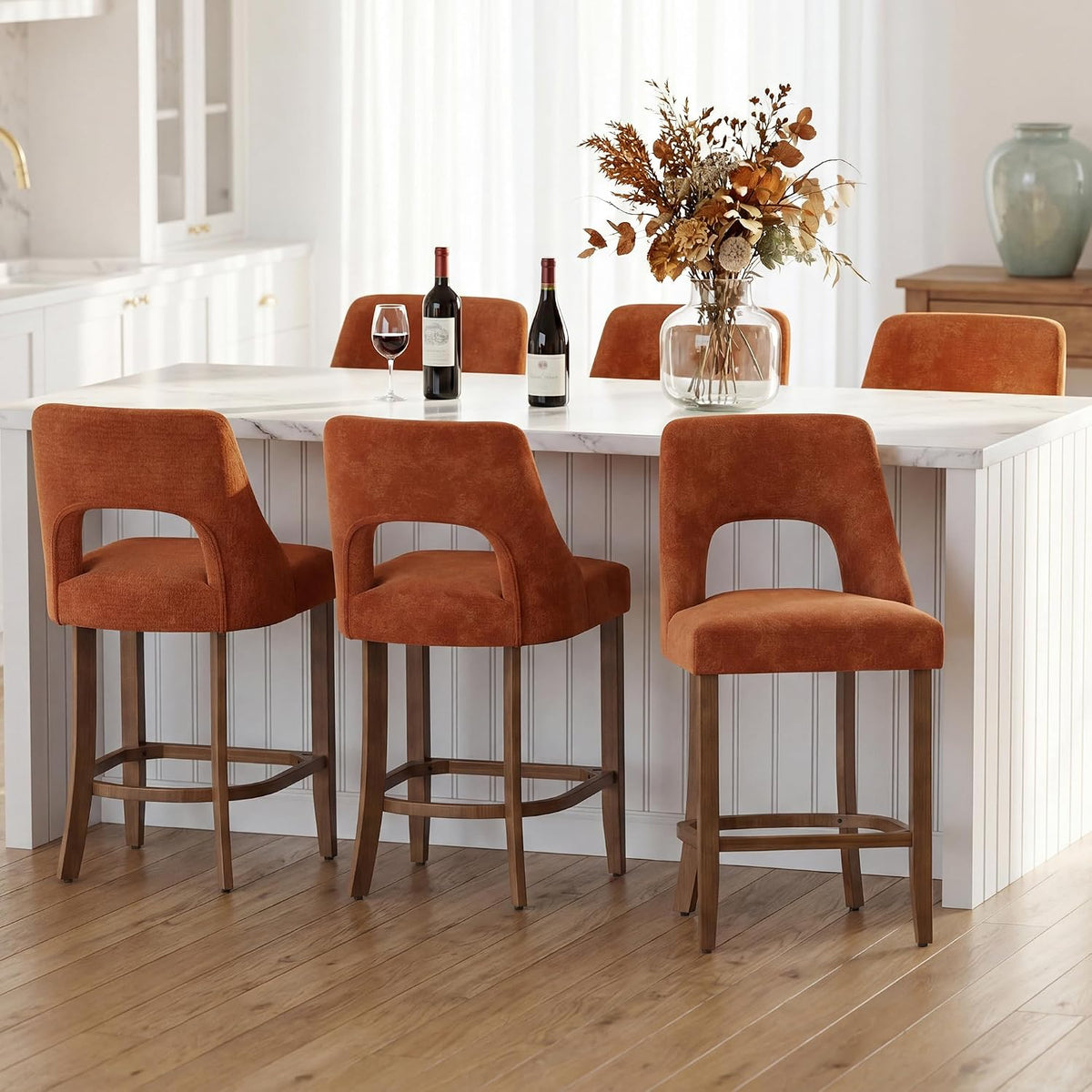 Three rust-orange upholstered counter stools arranged at a white kitchen island with wine and flowers on top.