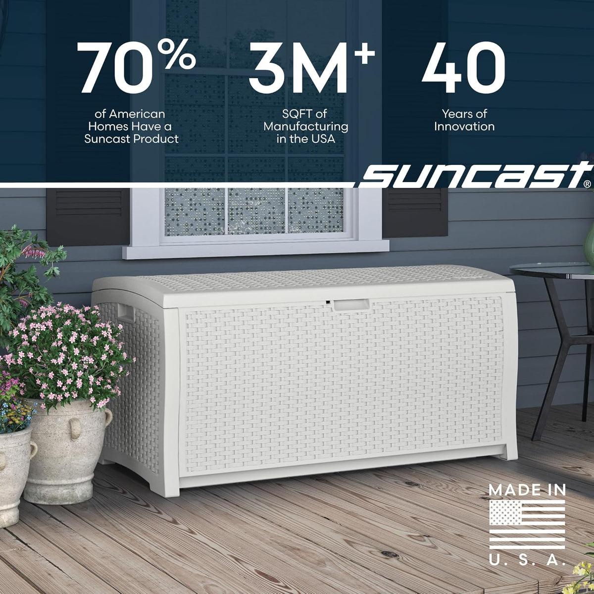 Front view of a white woven-effect deck box on a wood porch next to potted flowers and a window above.