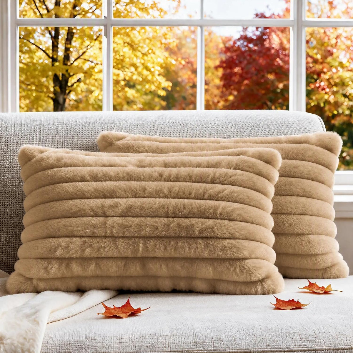 Two beige ribbed faux-fur pillows arranged on a sofa with scattered autumn leaves in front.
