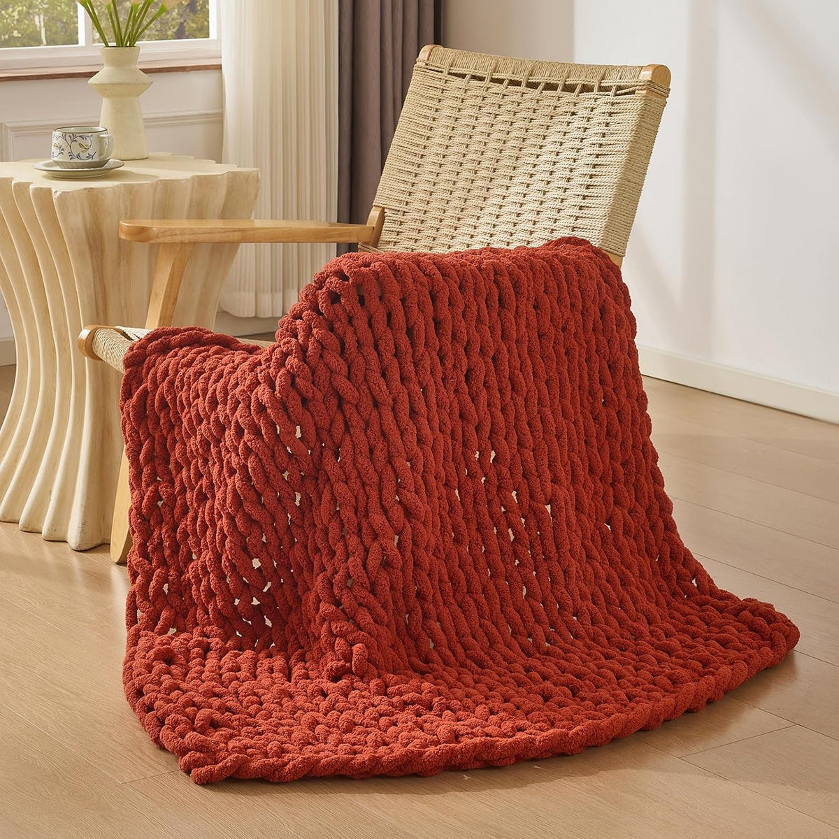Deep rust-red chunky knit throw draped over a woven-seat chair beside a side table.