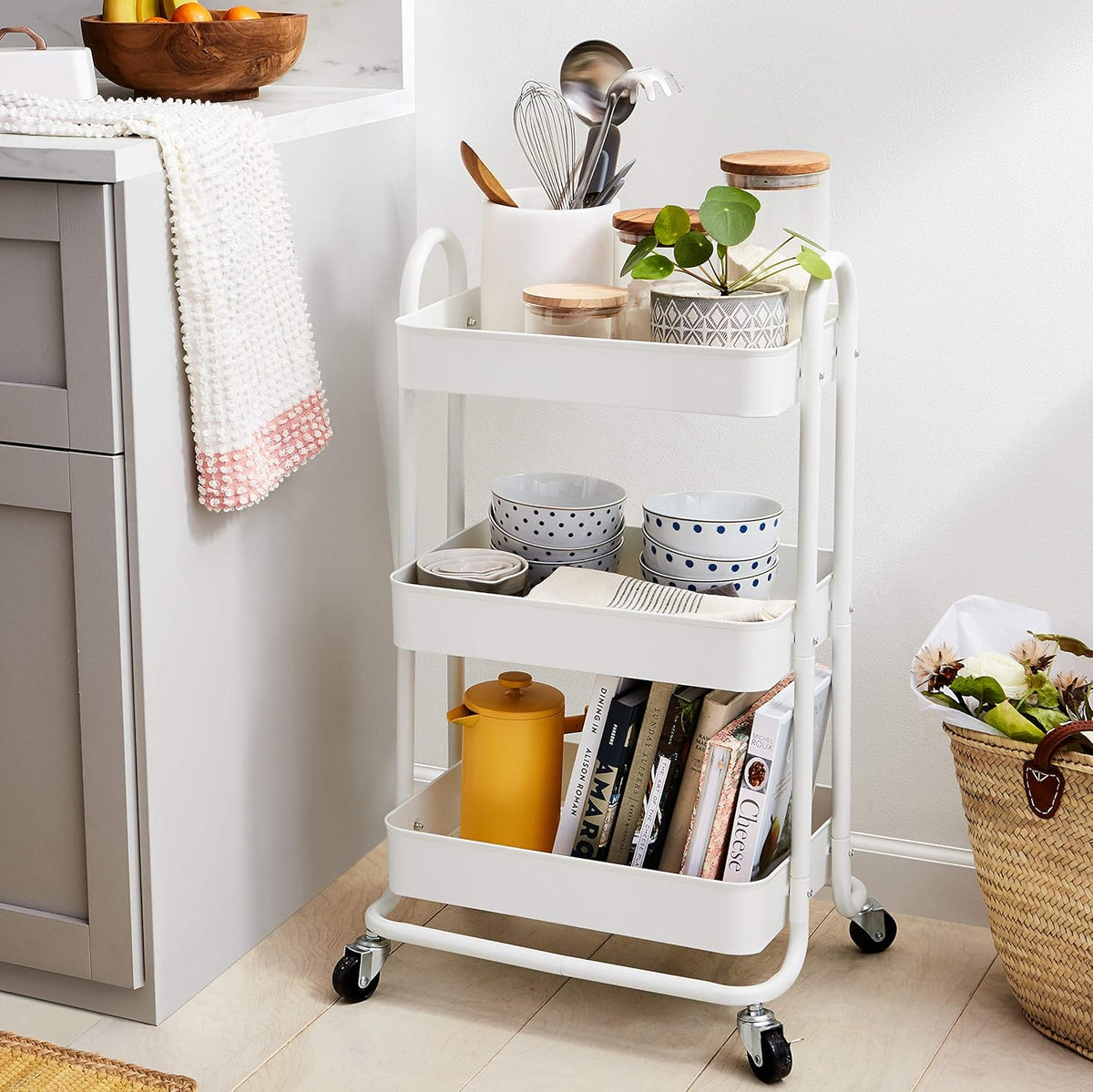 White three-tier rolling cart styled in a kitchen corner holding bowls, jars, books and a plant.