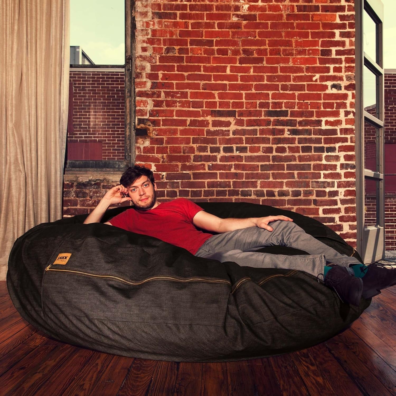 Man reclining on a large dark charcoal round bean bag in an industrial loft with exposed brick.