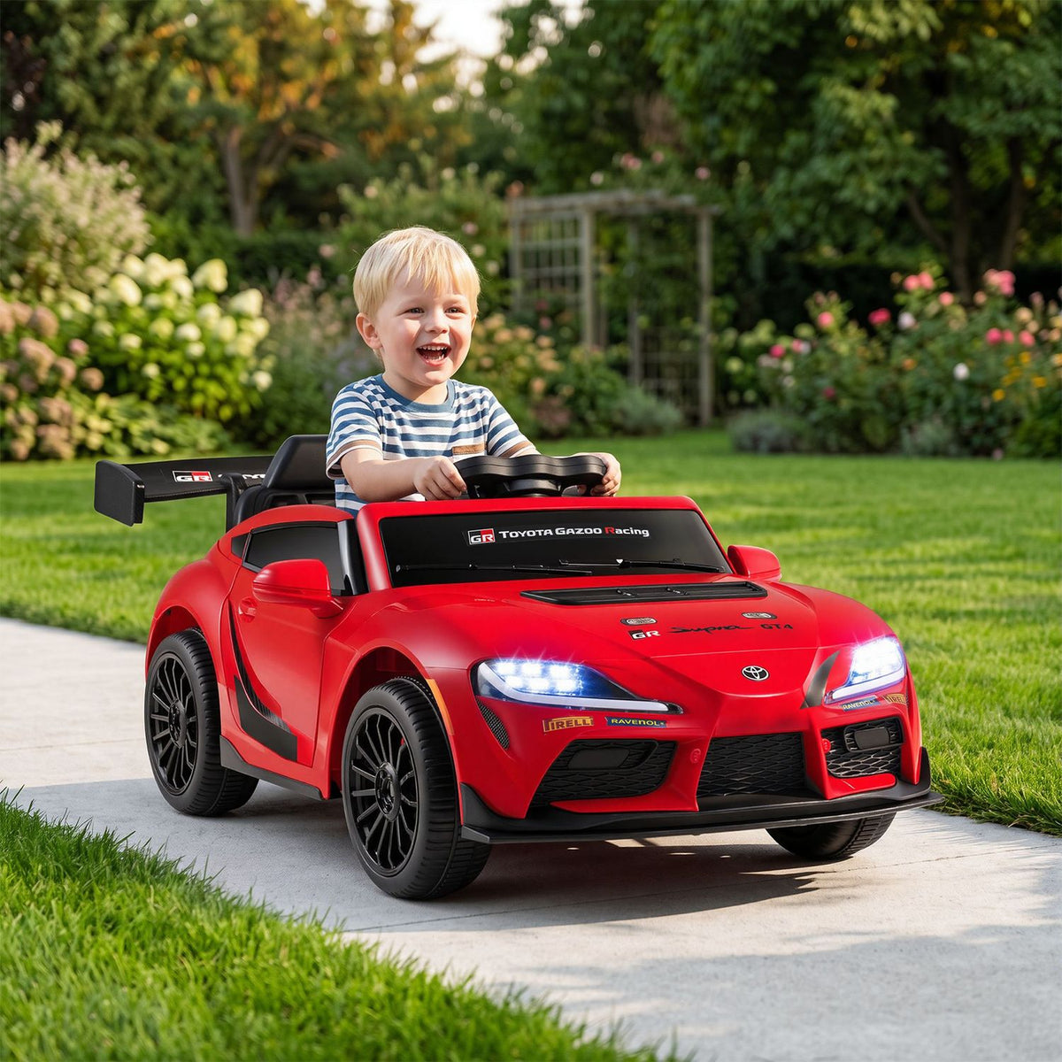 A child sits behind the wheel of a red ride-on sports car on a garden path.