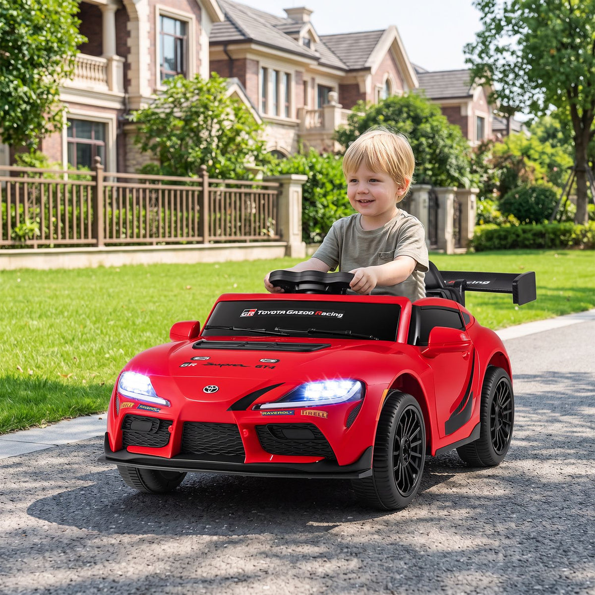 A child drives a red ride-on car across a paved path beside a lawn.