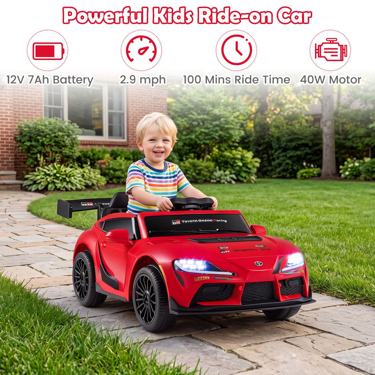 A child sits in a red ride-on car on a garden path, with the headlights lit.