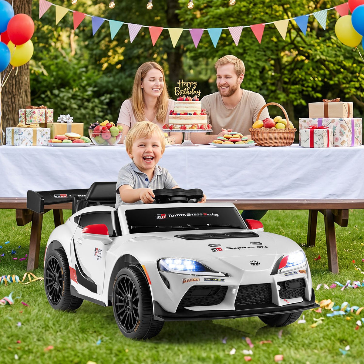 A child sits in a white ride-on sports car at an outdoor birthday party.