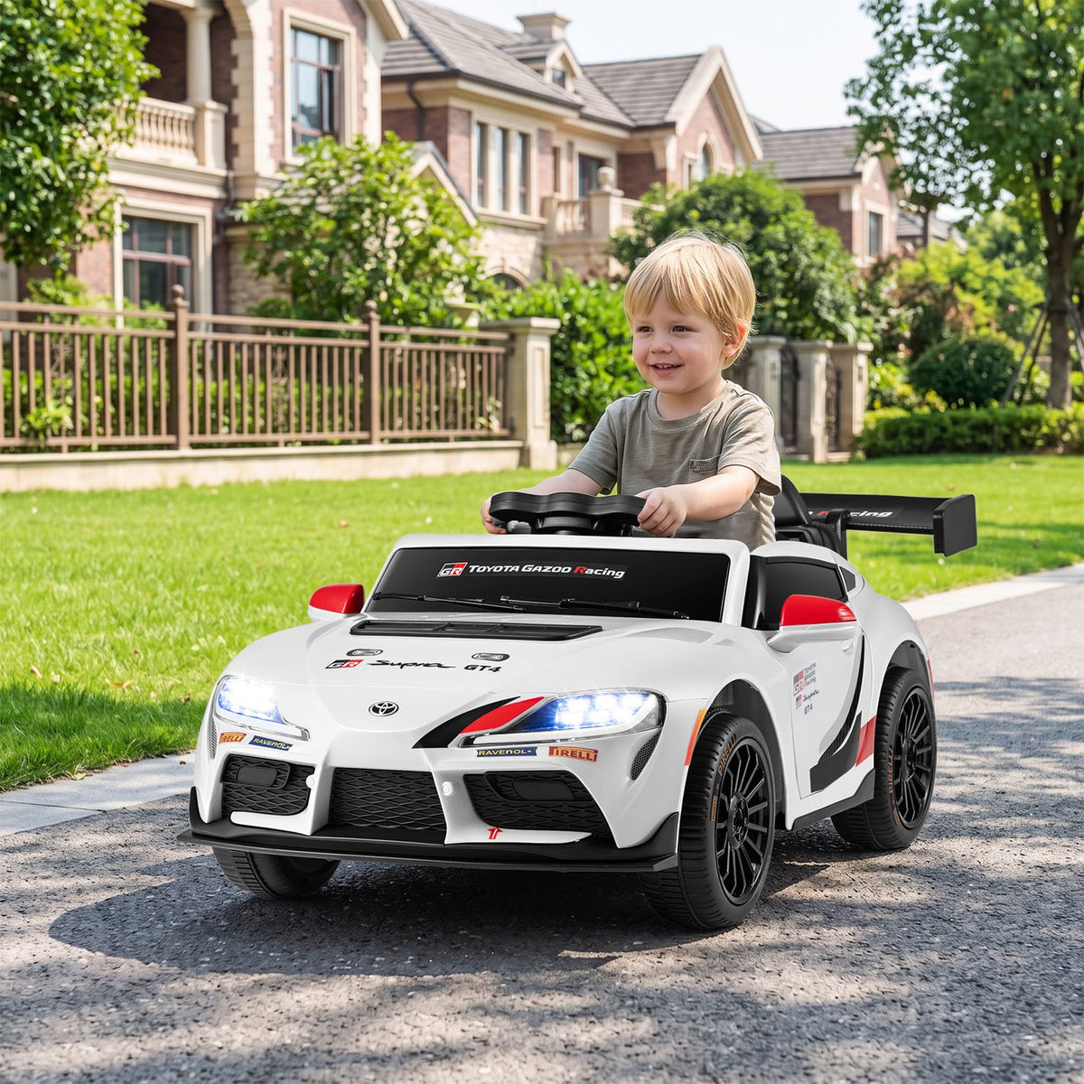 A child drives a white ride-on sports car along a path in a grassy neighborhood.