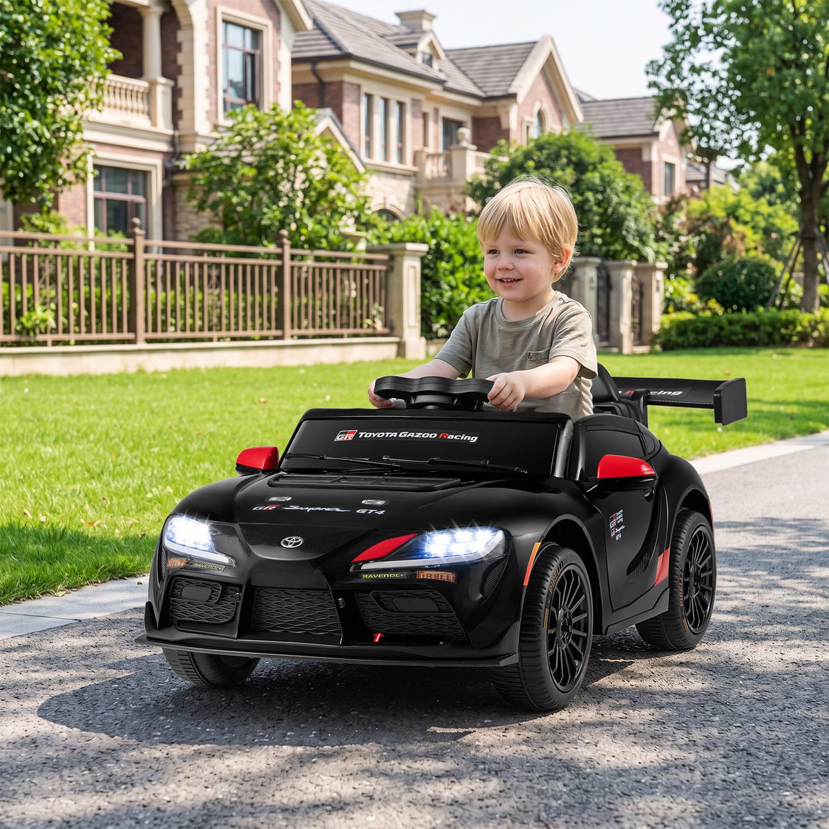 A child holds the wheel of a black ride-on car on a sunny residential walkway.