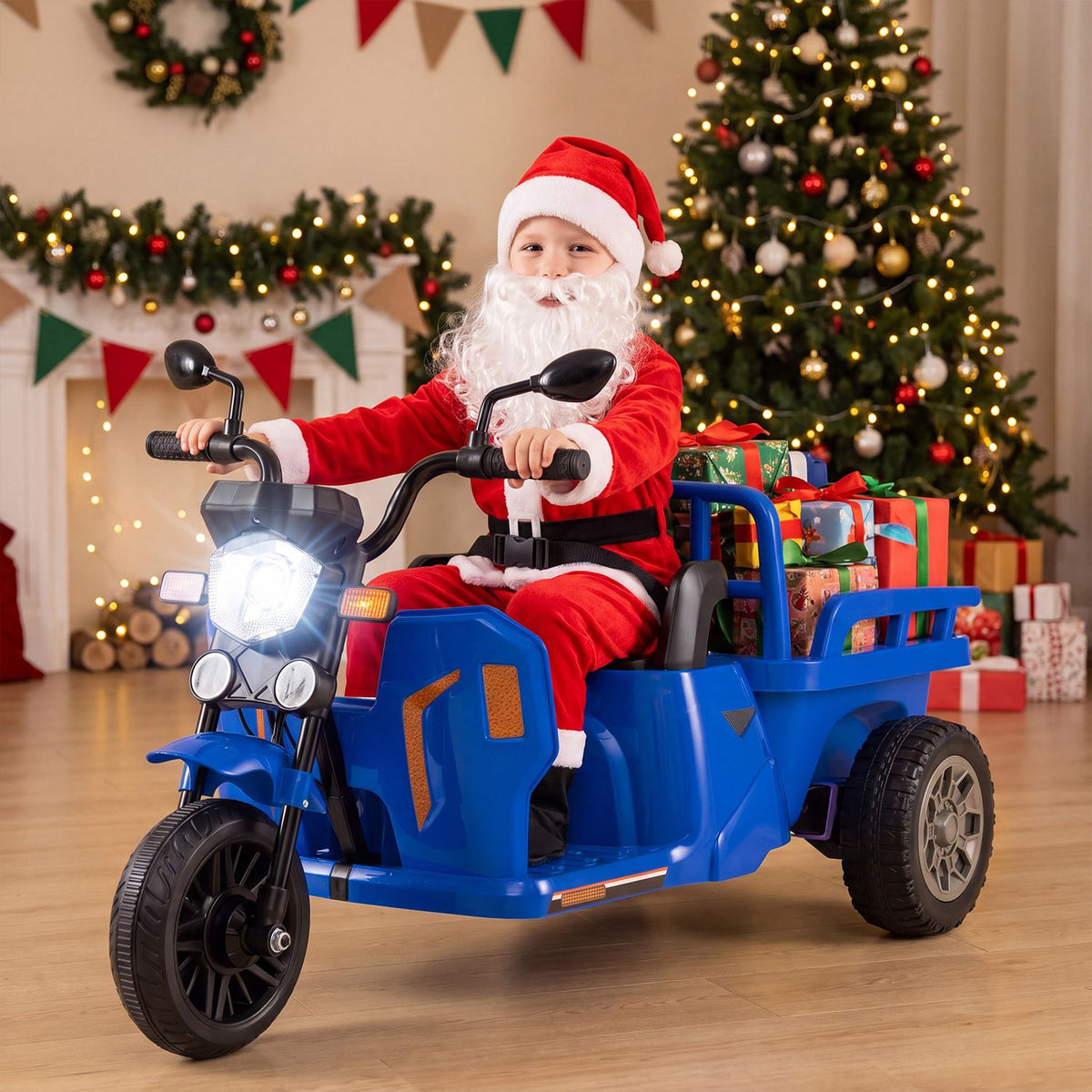 A child dressed as Santa rides a blue three-wheeled vehicle with a trailer beside a decorated Christmas tree.