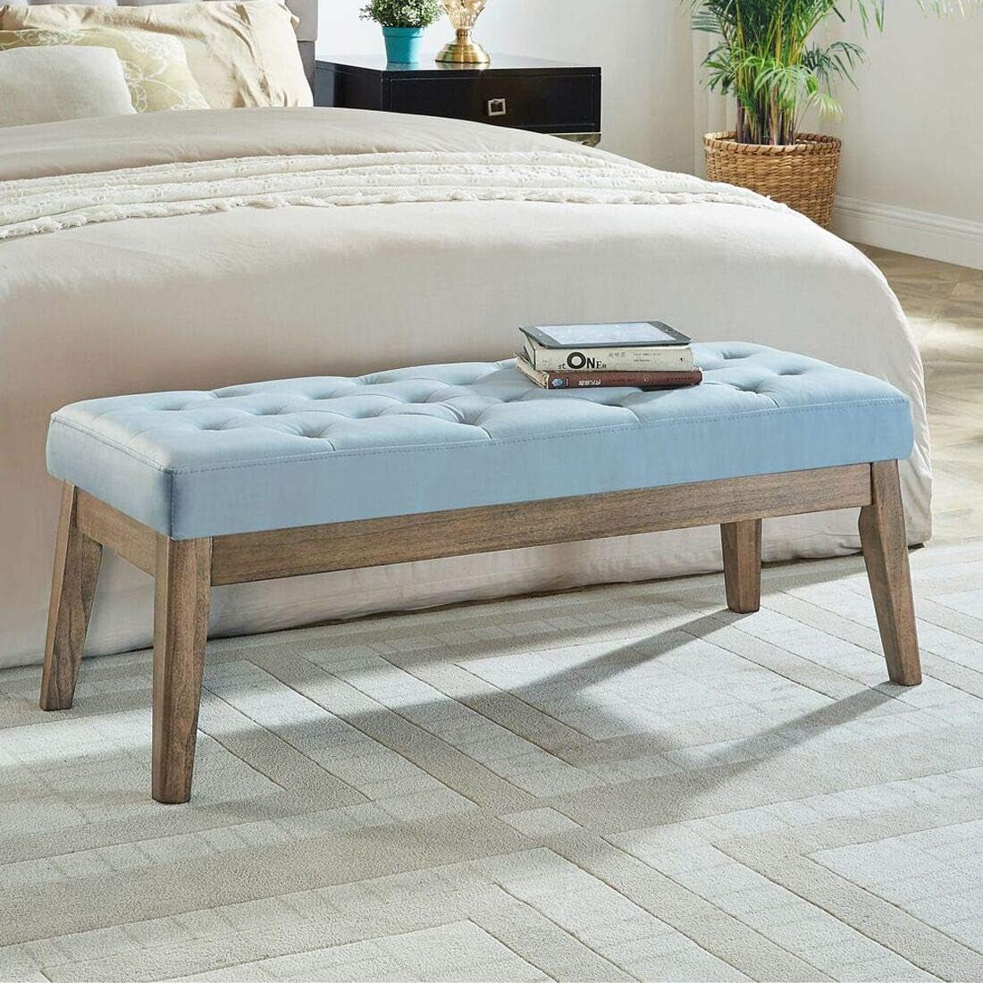 Light blue tufted bench at foot of bed with angled wood legs, a tablet and stacked books resting on the seat.