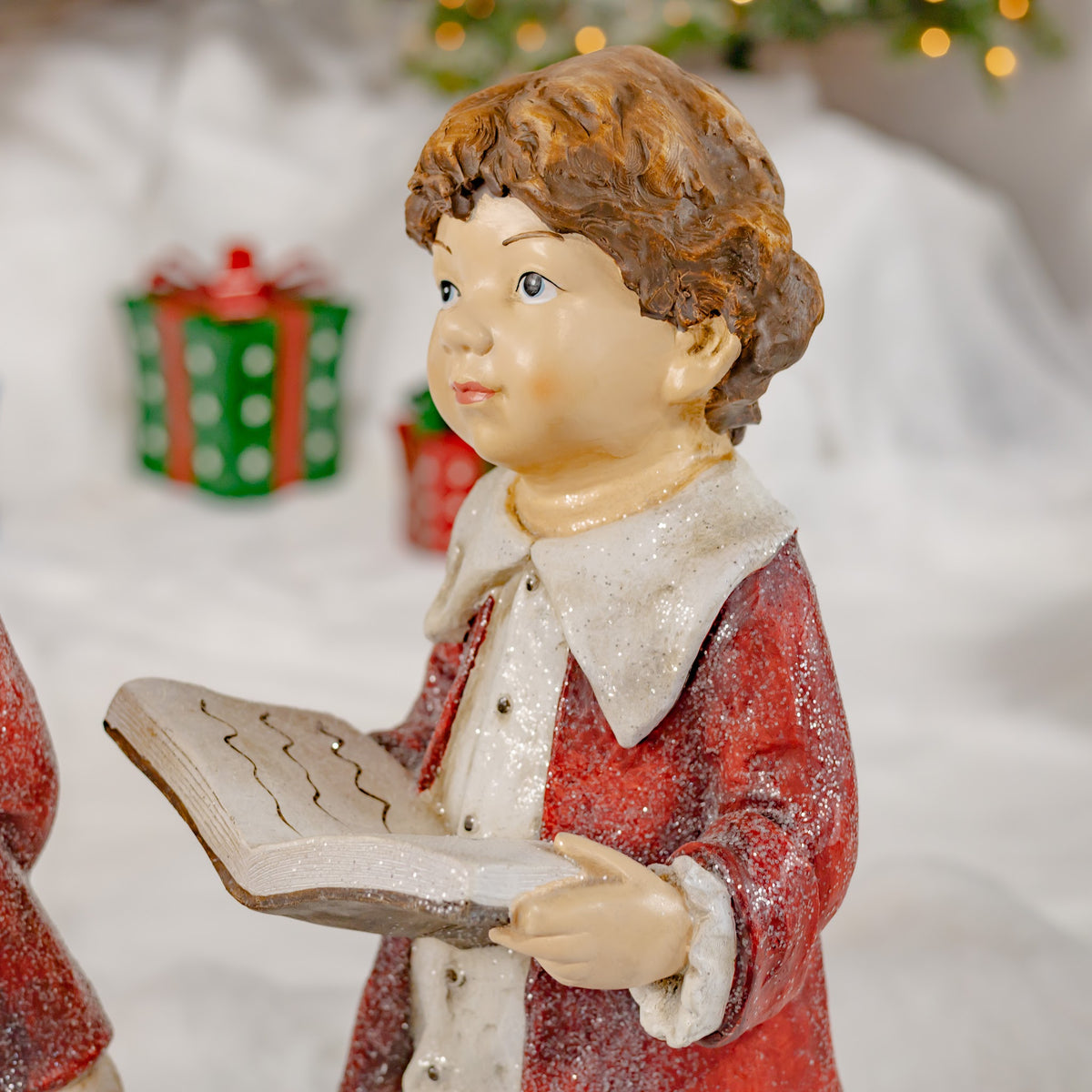 Close-up of a childlike figure in a red cloak holding an open book, with a second figure partly visible at the edge.