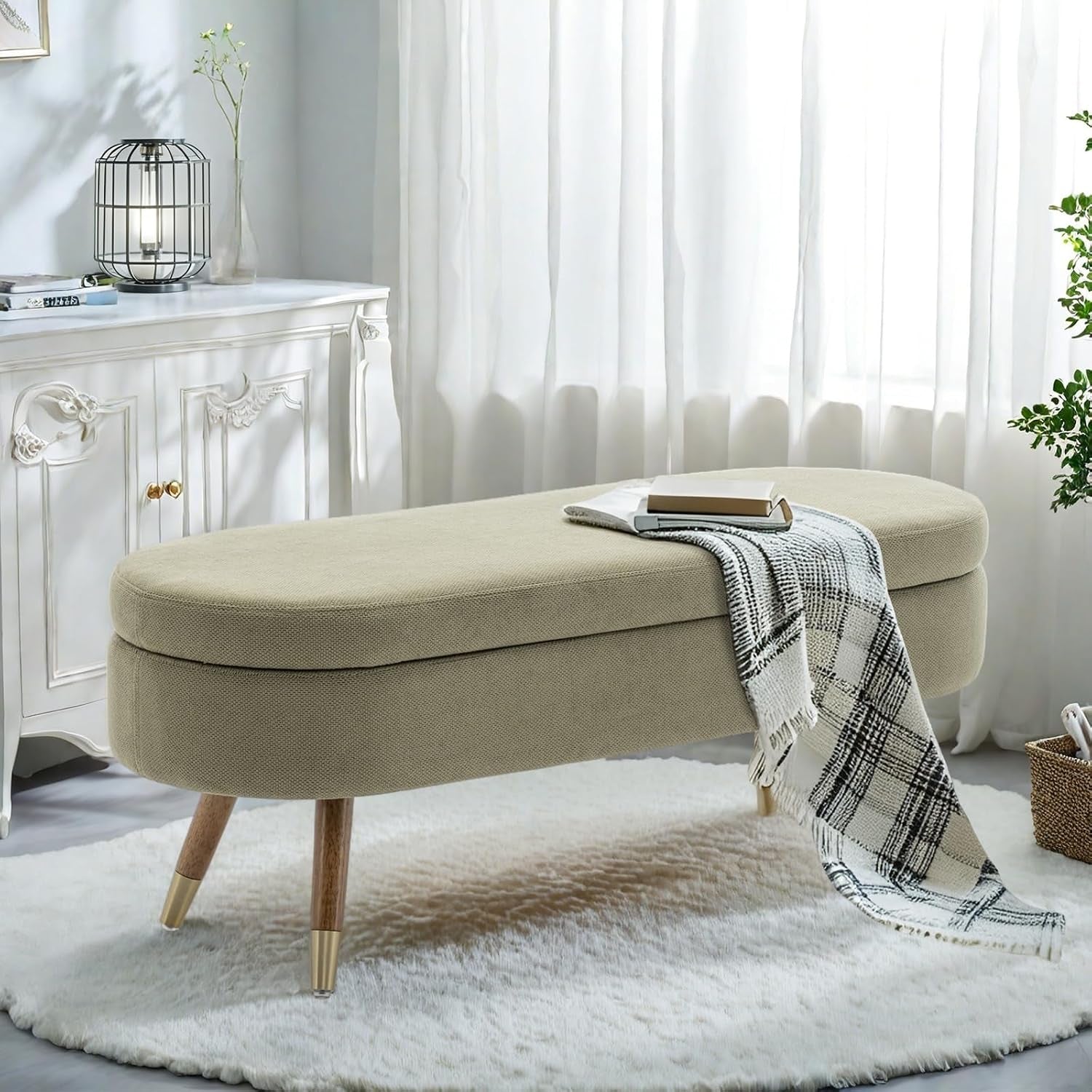 Long beige oval storage bench on wooden legs with gold feet placed on a round rug by a window.