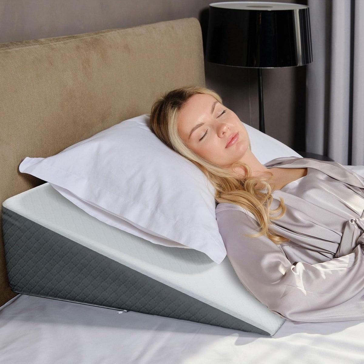 Woman reclining asleep on a white-top wedge pillow on a bed with a beige headboard, side view