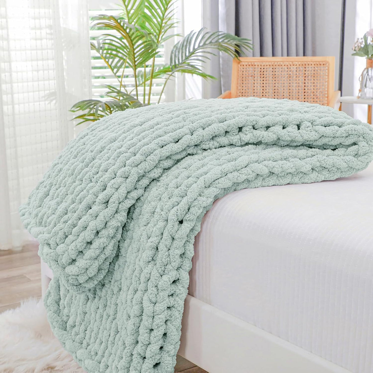 Folded mint-green chunky knit throw draped over a white bed edge with a cane-backed chair and plants behind.