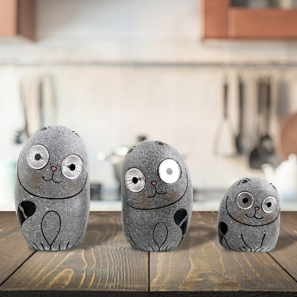 Three speckled gray cat ornaments with round eyes sit on a wooden table in a kitchen.