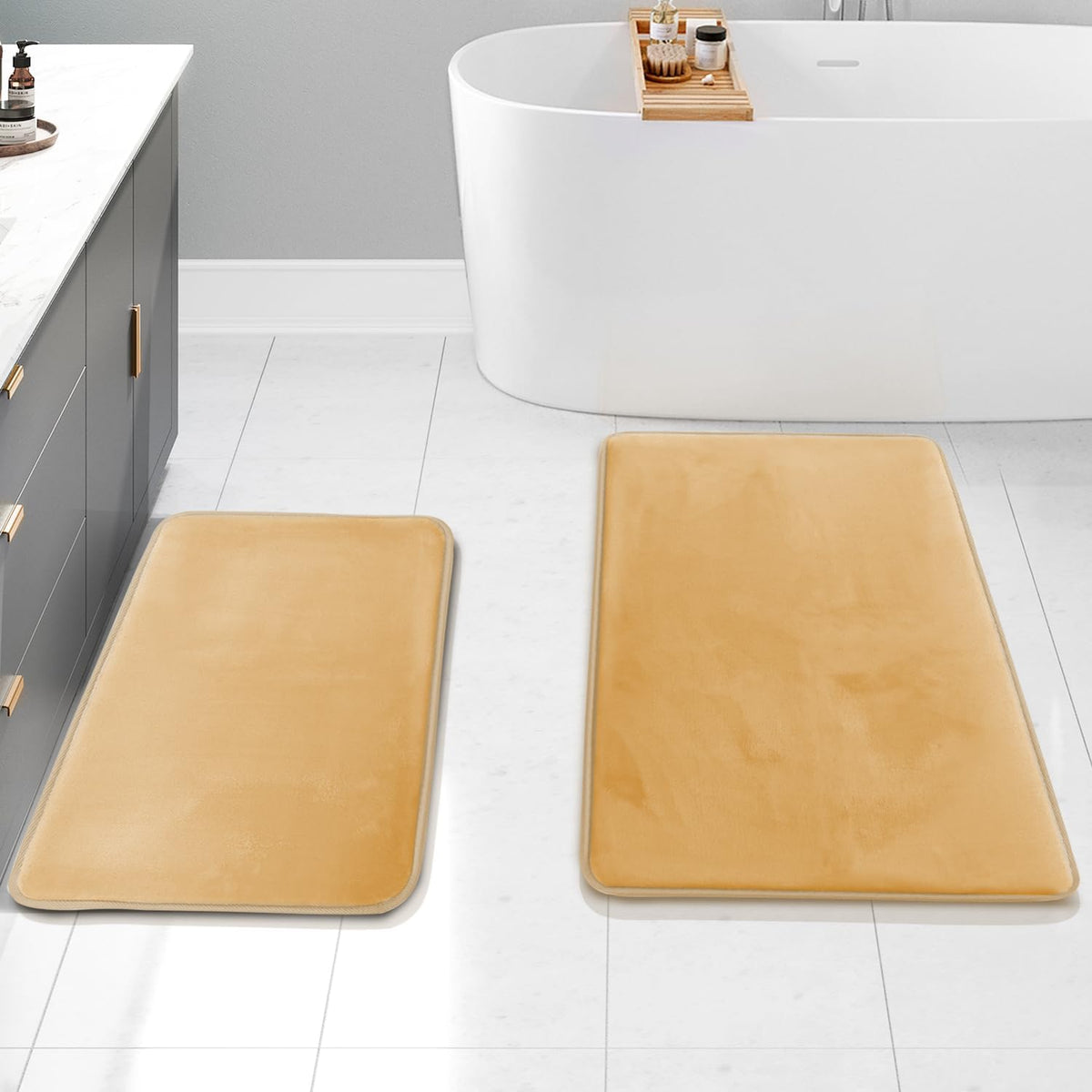 Bathroom scene with two camel/tan rectangular bath mats on a white tile floor near a tub and gray vanity.