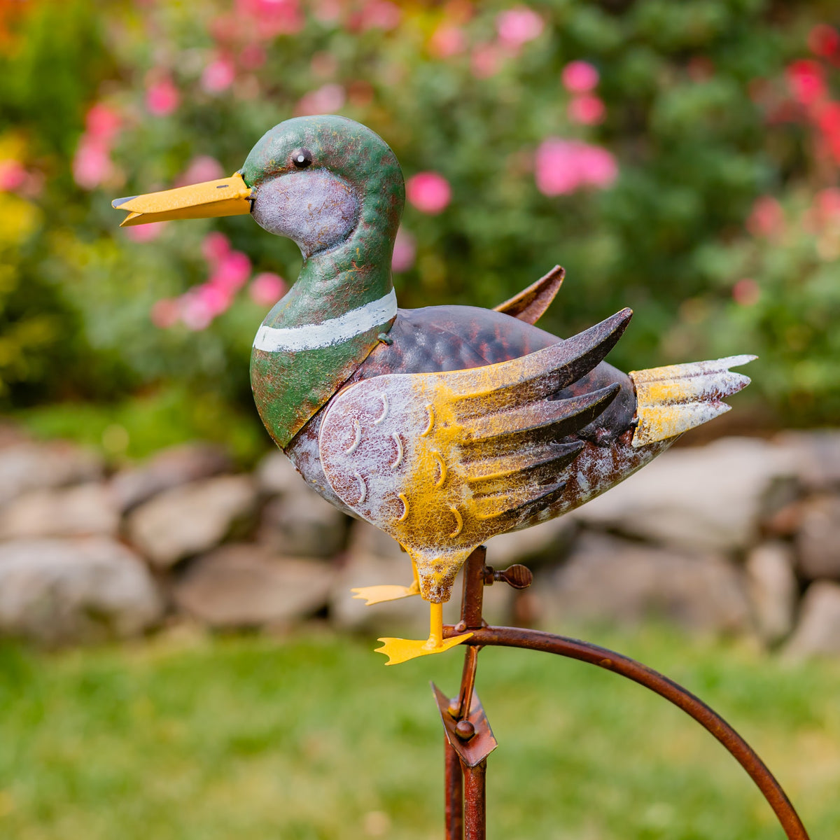 Side view of the painted duck figure and curved rocking mechanism against a blurred flower garden.
