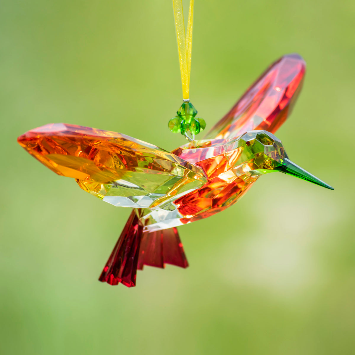 Faceted hummingbird ornament in orange, pink, and red tones, shown hanging against a blurred green background.