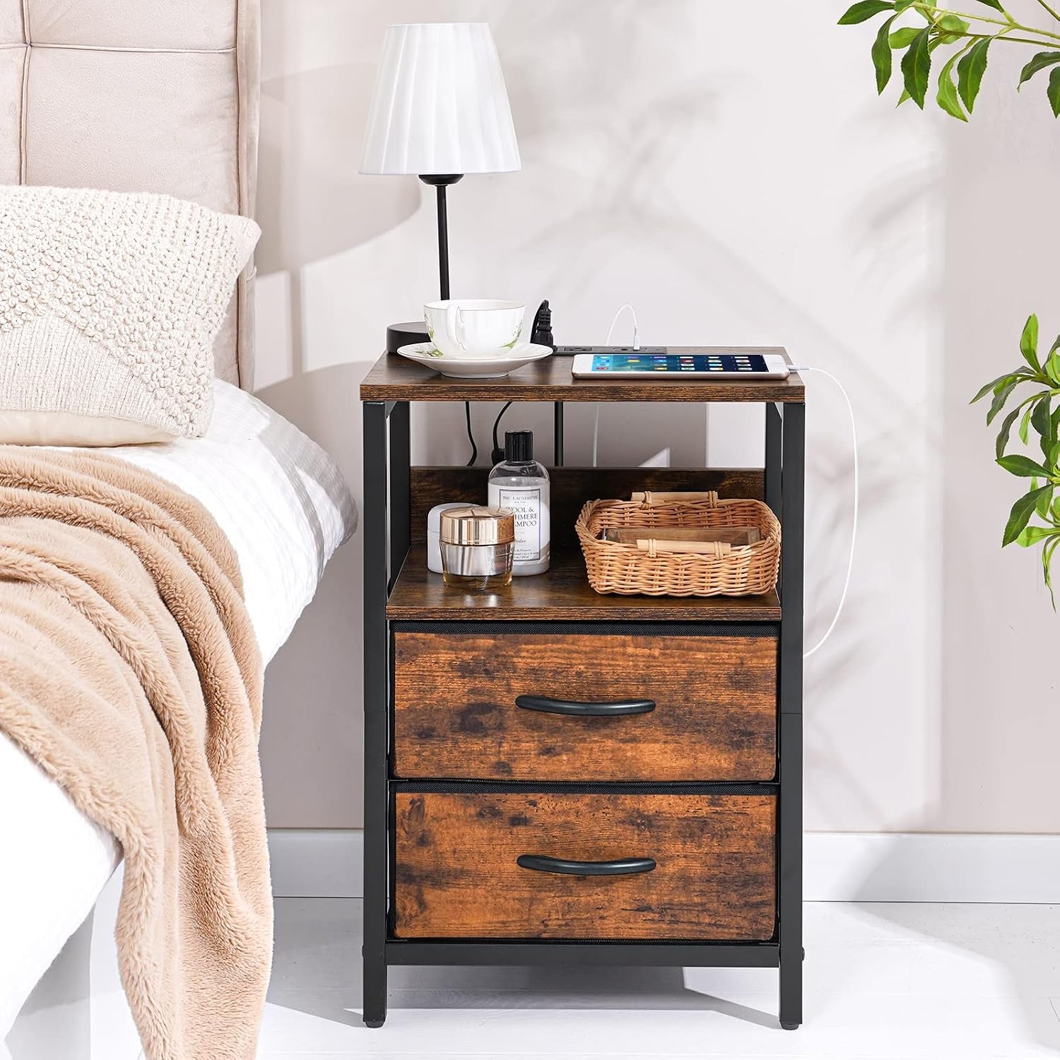 Nightstand placed beside a bed with lamp, basket and small items on shelves and a phone charging on top.