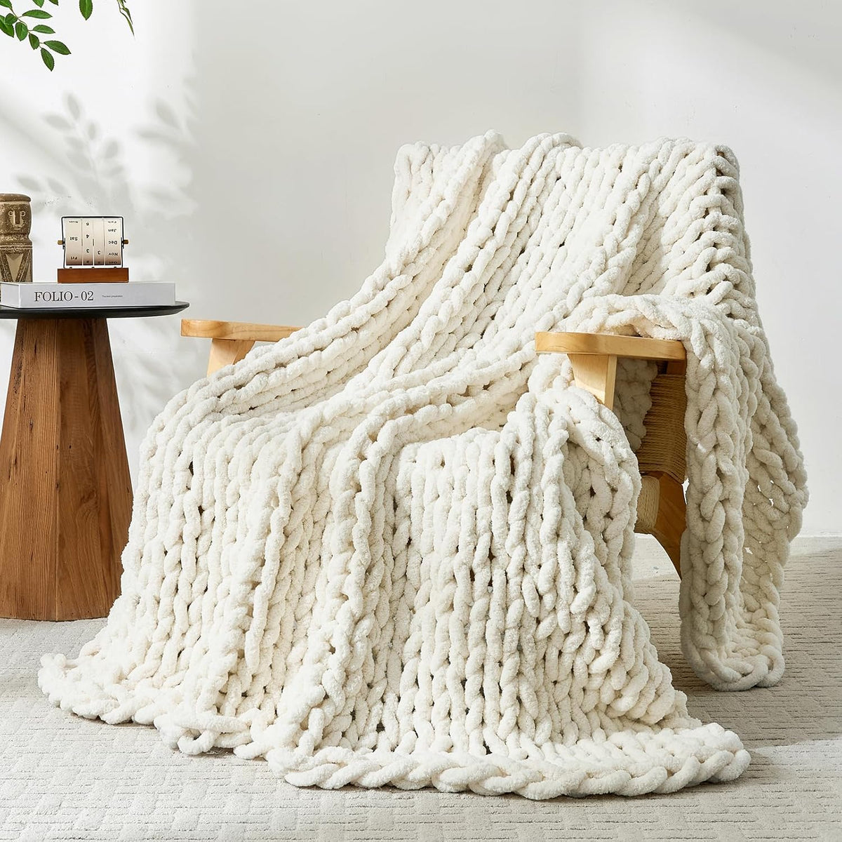 Beige chunky knit throw cascading from a low wood-framed lounge chair and pooling on a textured rug.