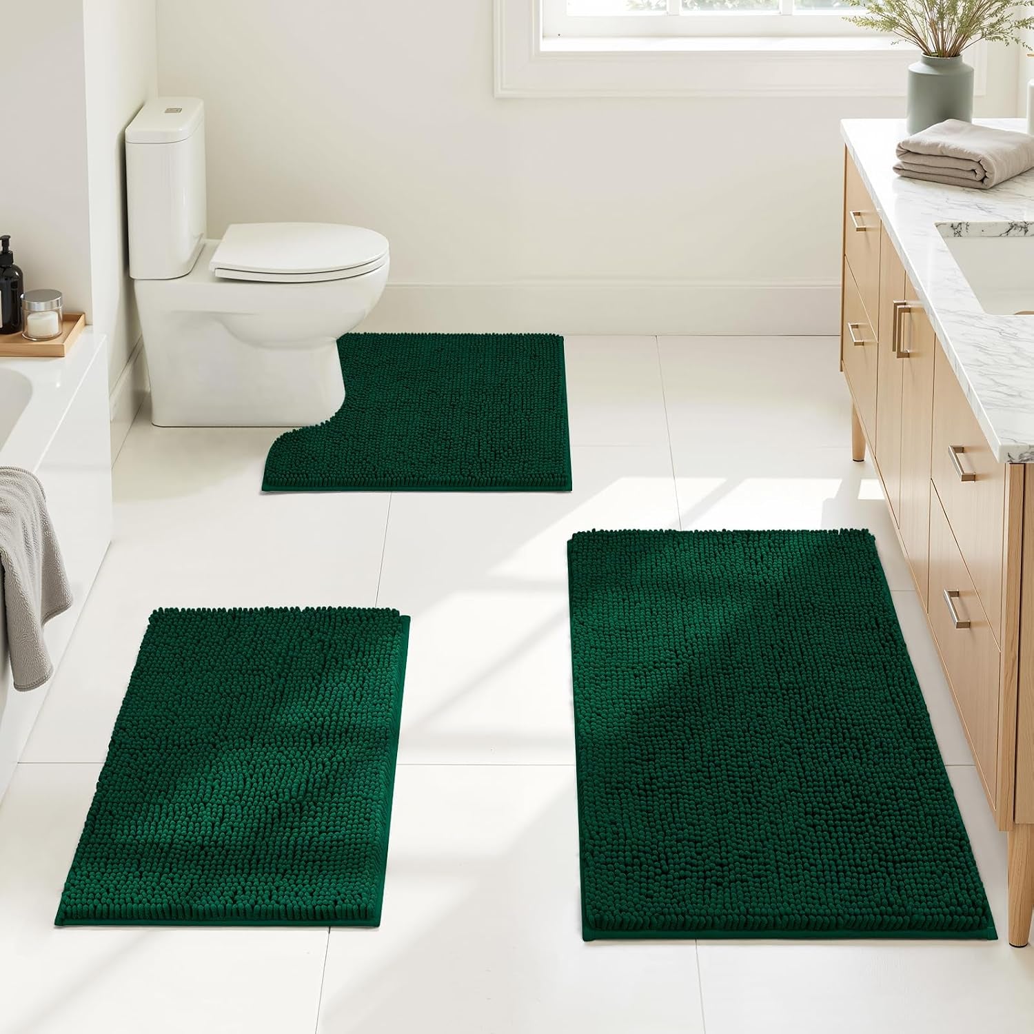Bathroom scene showing three dark green chenille rugs placed by bathtub, toilet (contoured), and sink cabinet on white tile