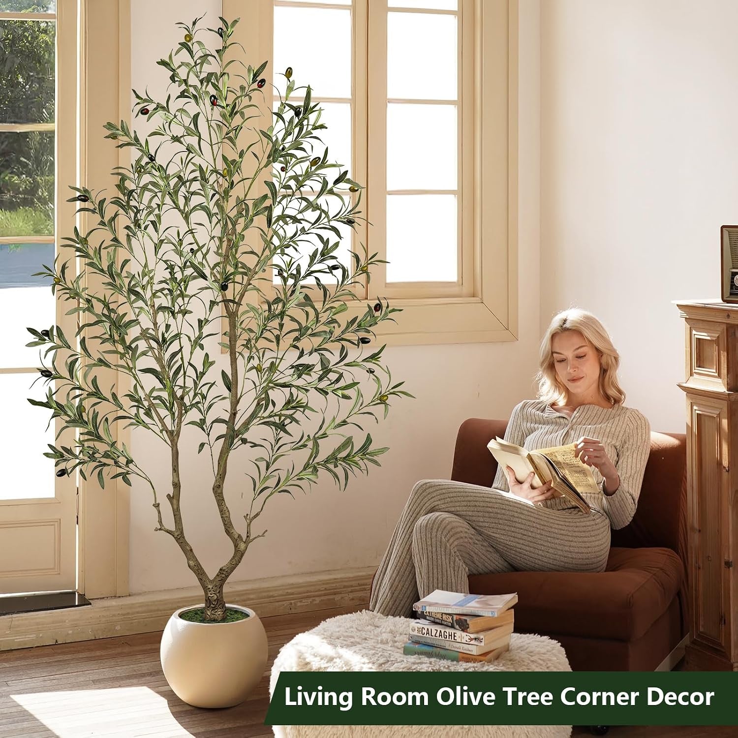 Olive tree beside tall windows in a living room while a woman reads on a nearby chair, books stacked on a footstool.