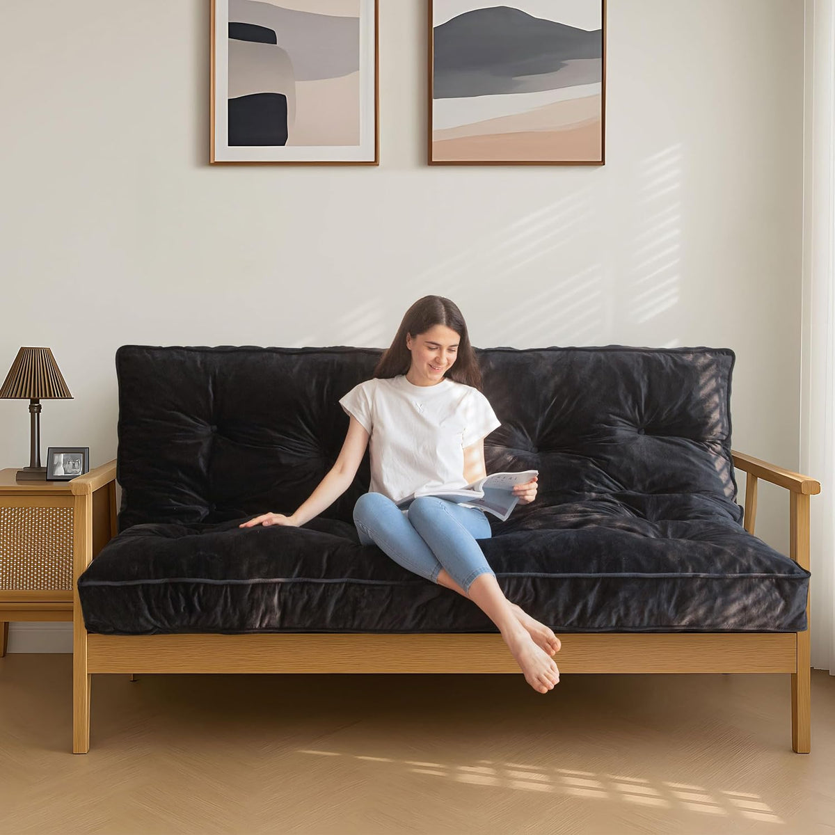 Black tufted futon mattress on a wooden frame shown with a woman reading beside framed wall art