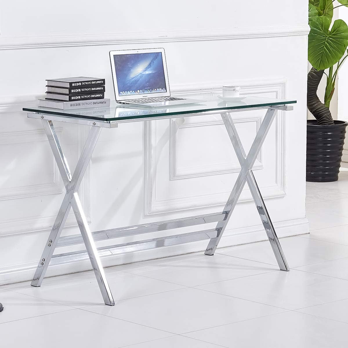 Clear tempered glass desk with chrome X-shaped legs, front-left view with laptop and stacked books