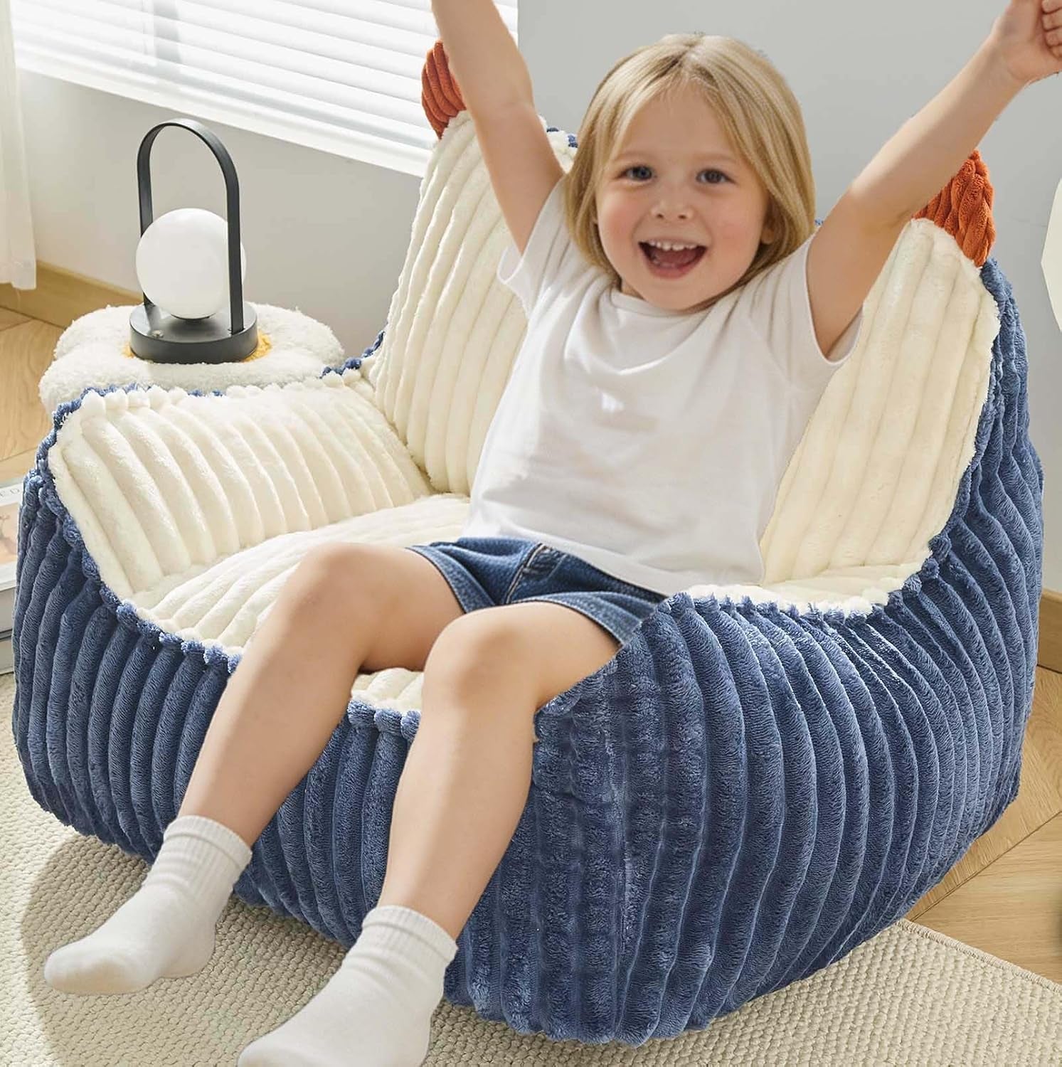 Happy child with arms raised sitting in a two-tone blue-and-cream ribbed round bean bag with small lamp nearby.