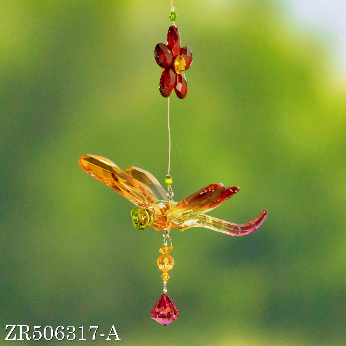 Amber dragonfly ornament with a red flower and pink dangling crystal against a blurred green background.