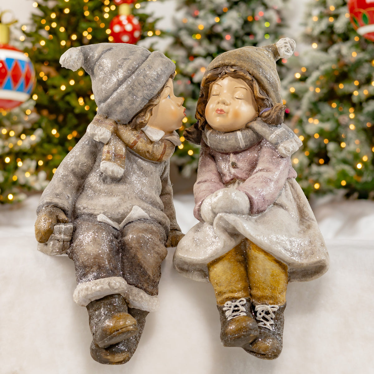 Two seated Christmas figures with dangling legs pose together in front of a lit tree.