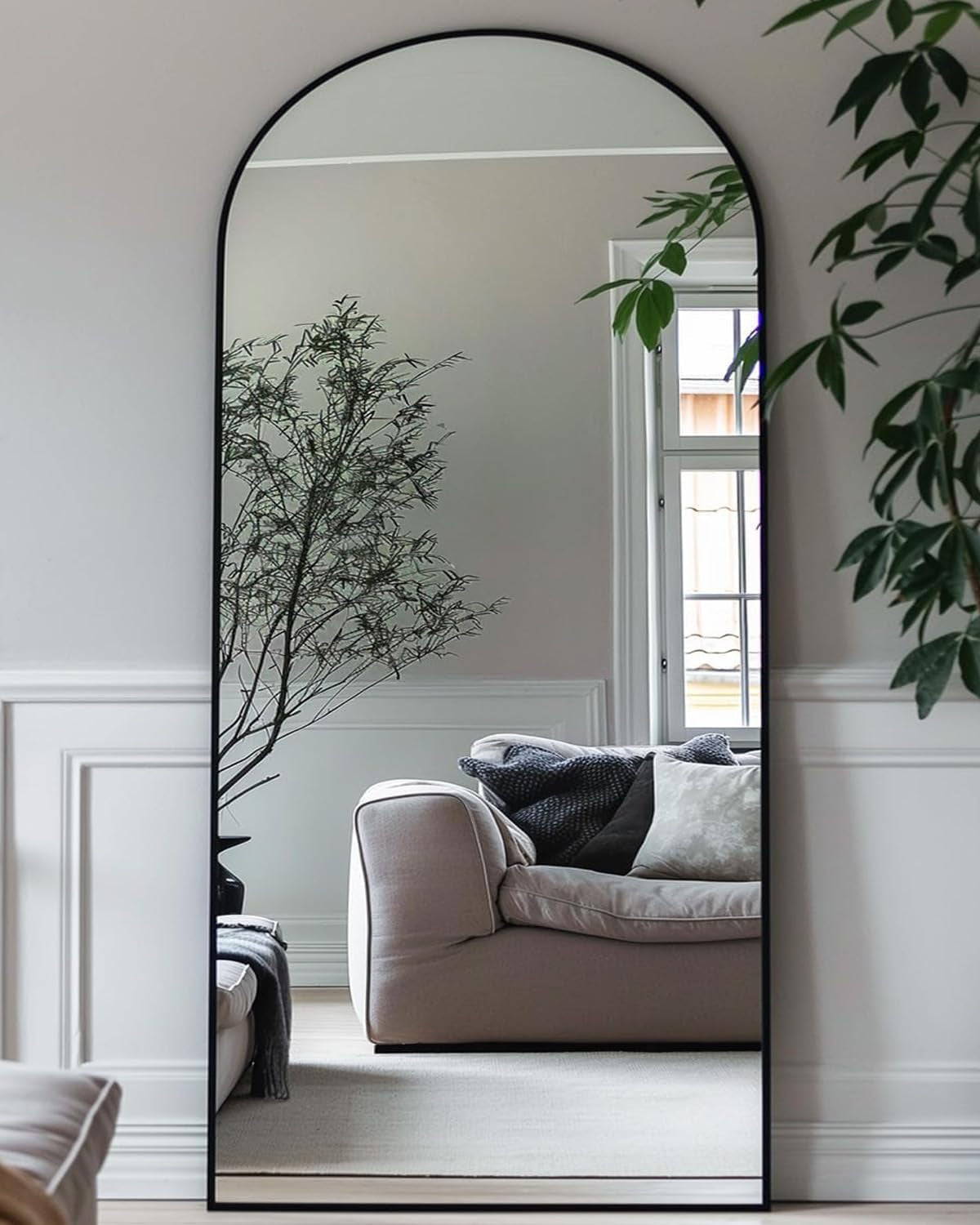 Tall arched mirror with black frame leaning against a paneled wall, reflecting a sofa, window, and indoor plants.