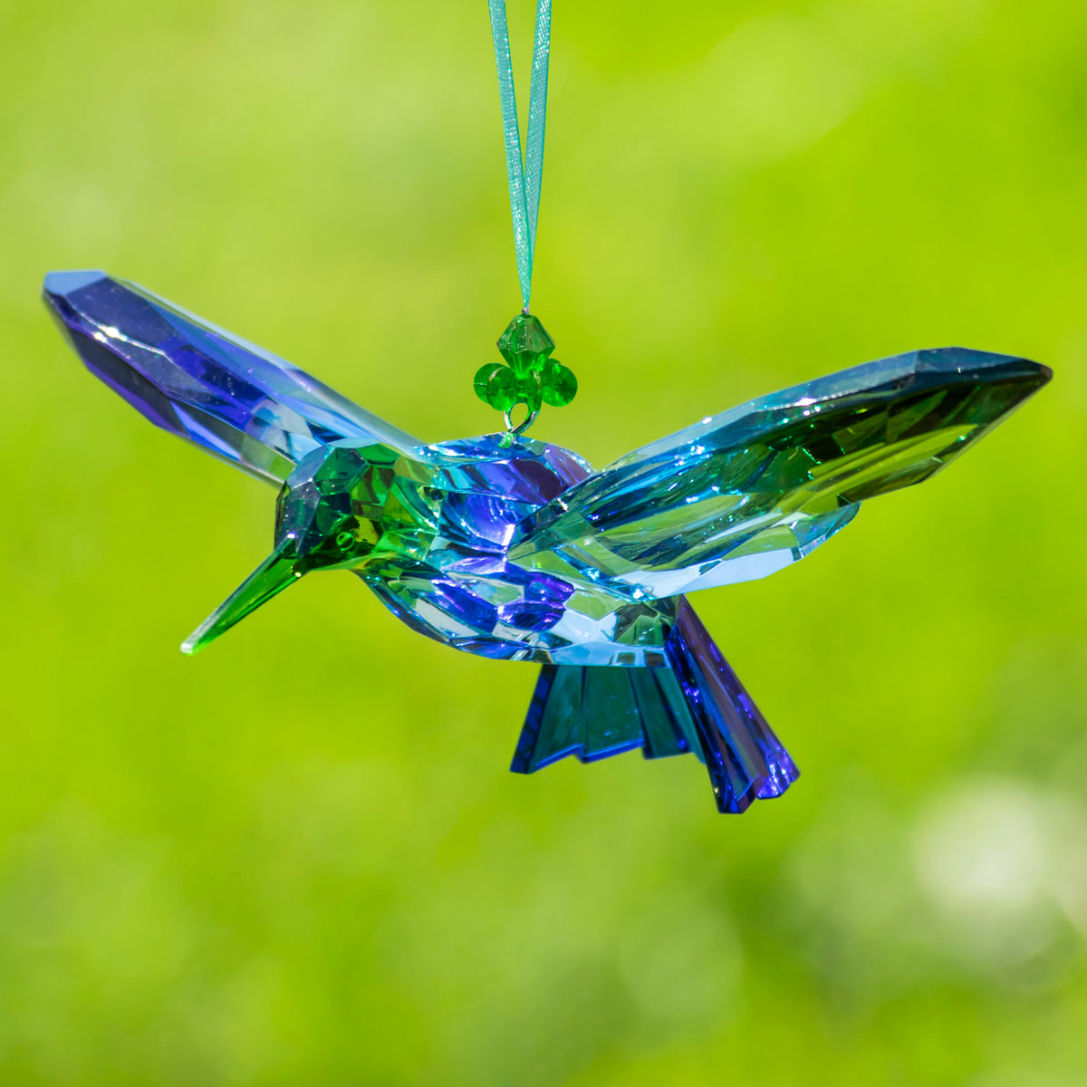 A blue and green faceted hummingbird ornament hangs with wings extended against a blurred green background.