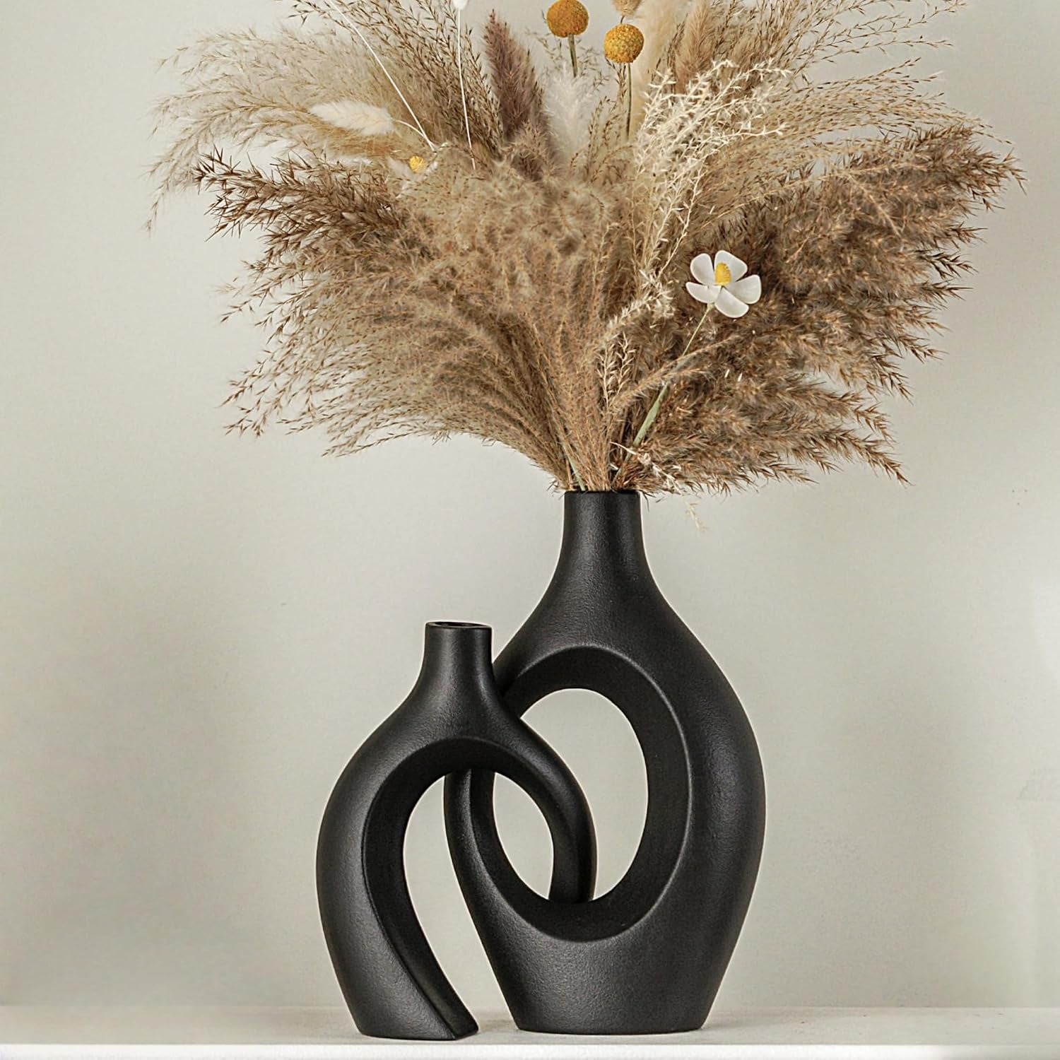 Two interlocking black ceramic vases holding dried grasses, shown on a low shelf against a pale wall.